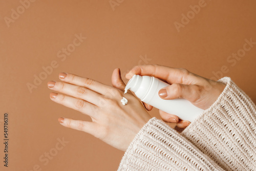 Woman holding cosmetic bottle and applying moisturizing cream on her hands, pastel brown background, skincare, aesthetic concept