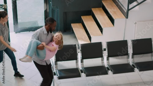 Worried man bringing unconscious girl in hospital lobby, kid fainting and parents asking physician for help. Senior medic and nurse consulting young sick girl in health center. Handheld shot.