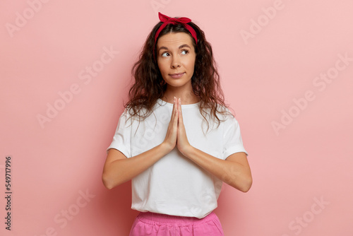 Young pretty woman begging to buy her new toy, she holds palms together and look aside, isolated over pink wall photo