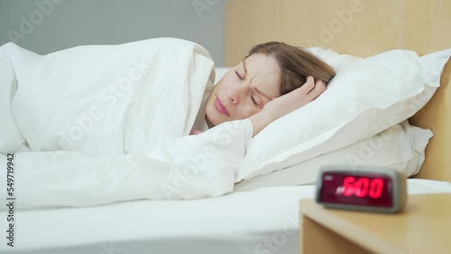 Close up portrait of sleeping woman in bed at home wakes up from the loud sound of the alarm clock on table and ignoring turns away to sleep covering with blanket in the bedroom Early morning concept