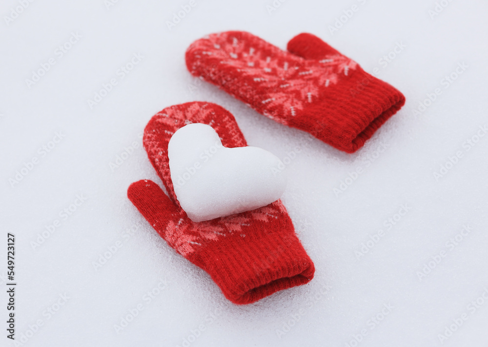 A snowy heart lies on red, knitted mittens in the snow. Selective focus