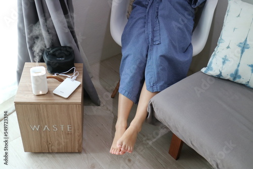 A person in a bathrobe sits on a chair with ankles exposed, next to a scent machine in use that emits the smoke of essential oils