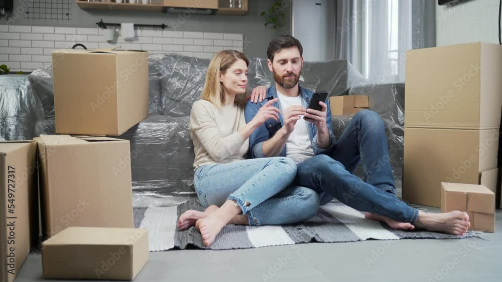 Smiling married couple plan apartment decoration browsing online stores using smartphone sitting in a living room of their new home with a lot of boxes in the background Moving relocation concept Smiling married couple plan apartment decoration browsing online stores using smartphone sitting in a living room of their new home with a lot of boxes in the background Moving relocation concept