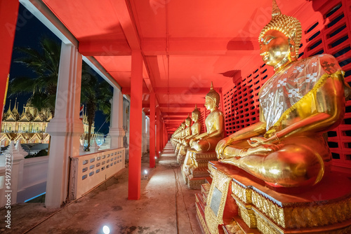 Several golden statues of Buddha statues in meditation posture are lined up in a corridor and spotlight on every statues in front of the red wall.