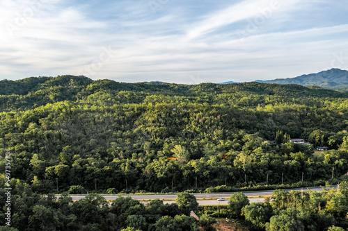 Aerial landscape of 4 lane road with residential area, forest, mountain, blue sky and sunlight with high angle view of main road from Drone shot in Tak province, northern countryside of Thailand.