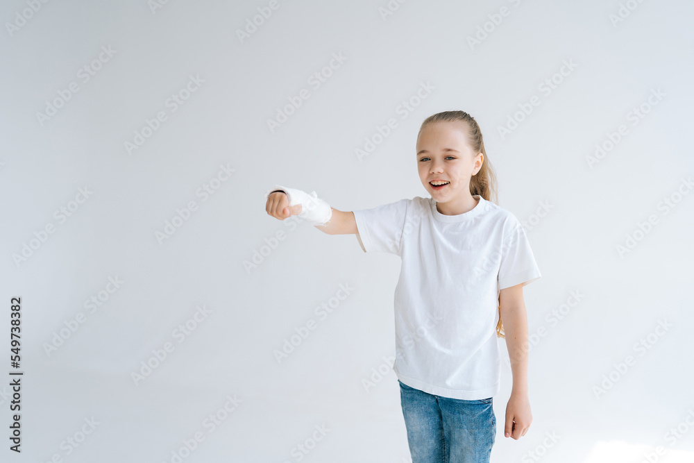 Portrait of positive little girl with broken hand wrapped in white plaster bandage playing gesturing with injured wrist on light isolated background. Concept of child insurance and healthcare. Portrait of positive little girl with broken hand wrapped in white plaster bandage playing gesturing with injured wrist on light isolated background. Concept of child insurance and healthcare.