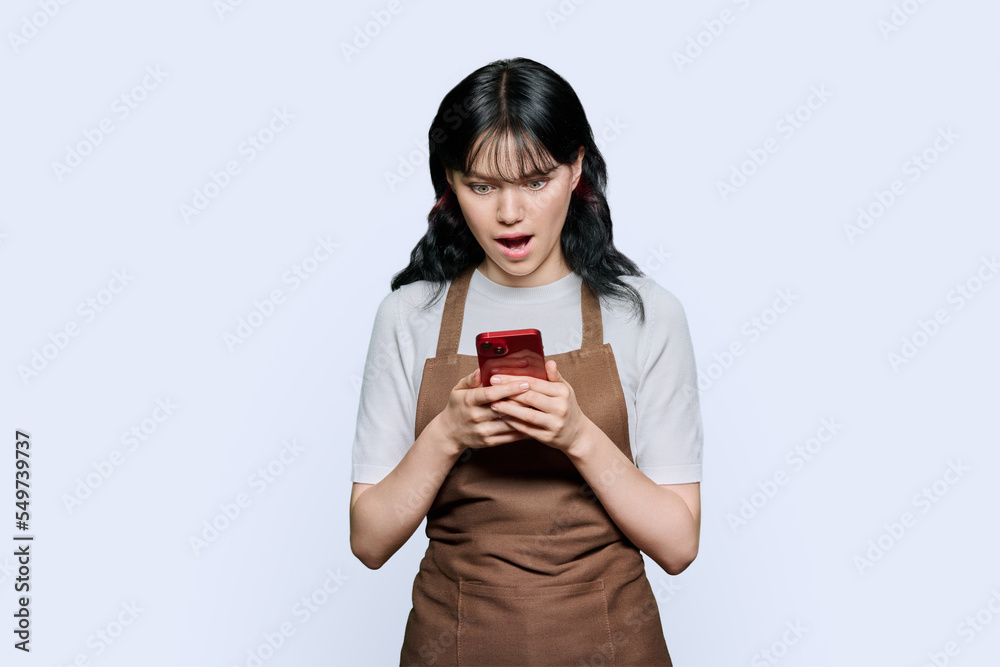 Surprised young woman in apron looking at smartphone screen