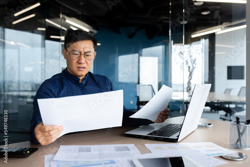 Asian businessman not satisfied with achievement results, boss looking at documents and upset, man in glasses and shirt working inside office with paperwork using laptop.