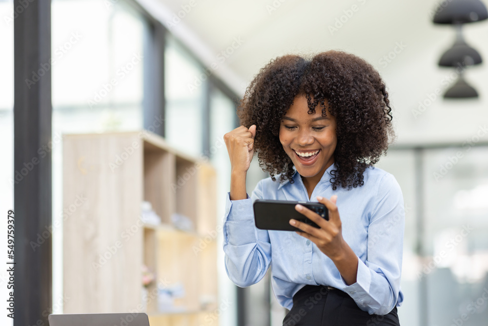 Fototapeta premium Successful and exciting Young african american business woman looking at the phone screen, african woman excited to win on her smartphone at work.