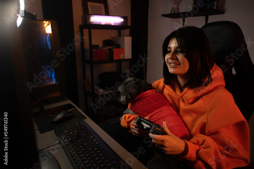 gamer girl in orange sweatshirt plays video games on her computer in her set-up with her pet on her lap.