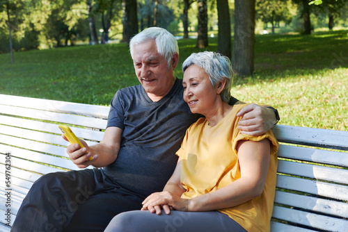 Senior spouses sit on bench in summer park with smartphone © Alex Tihonov