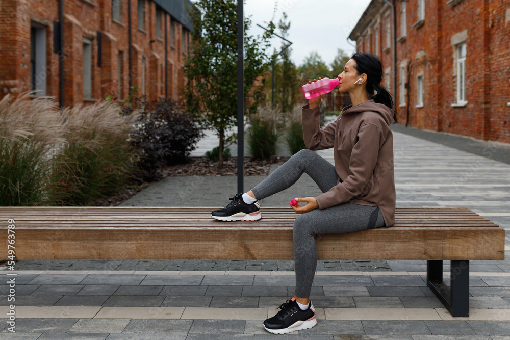 Sports woman in earphones drinking water after jogging on the bench