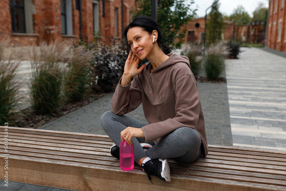 Beautiful fitness woman with sports bottle of water in hands enjoying motivational music in earphones while sitting on bench after workout outdoors