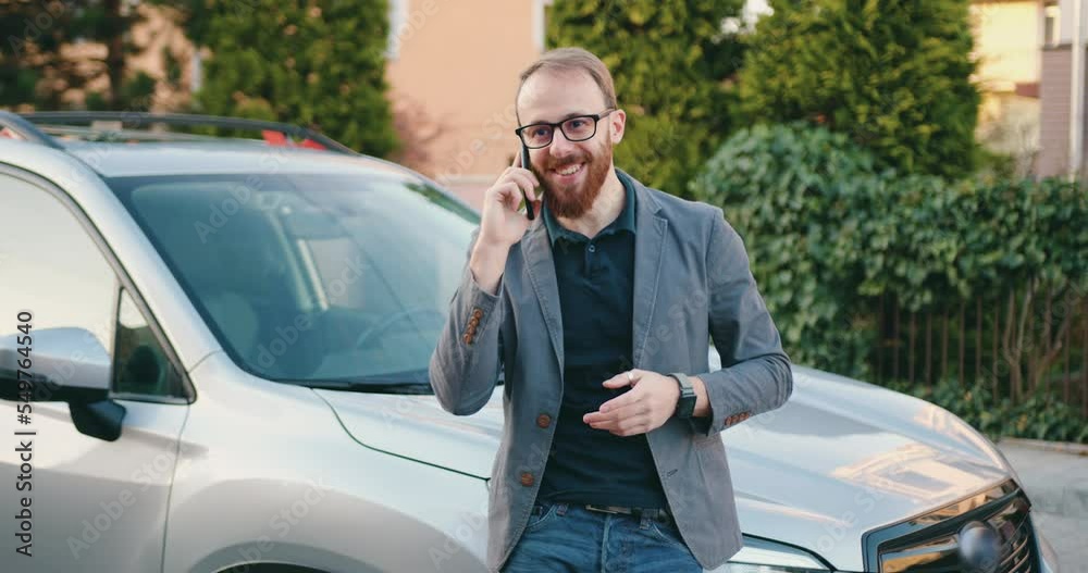 Smiling businessman talking on cell phone answering business call when leans on her car which is parked by area house. Outdoors Smiling businessman talking on cell phone answering business call when leans on her car which is parked by area house. Outdoors
