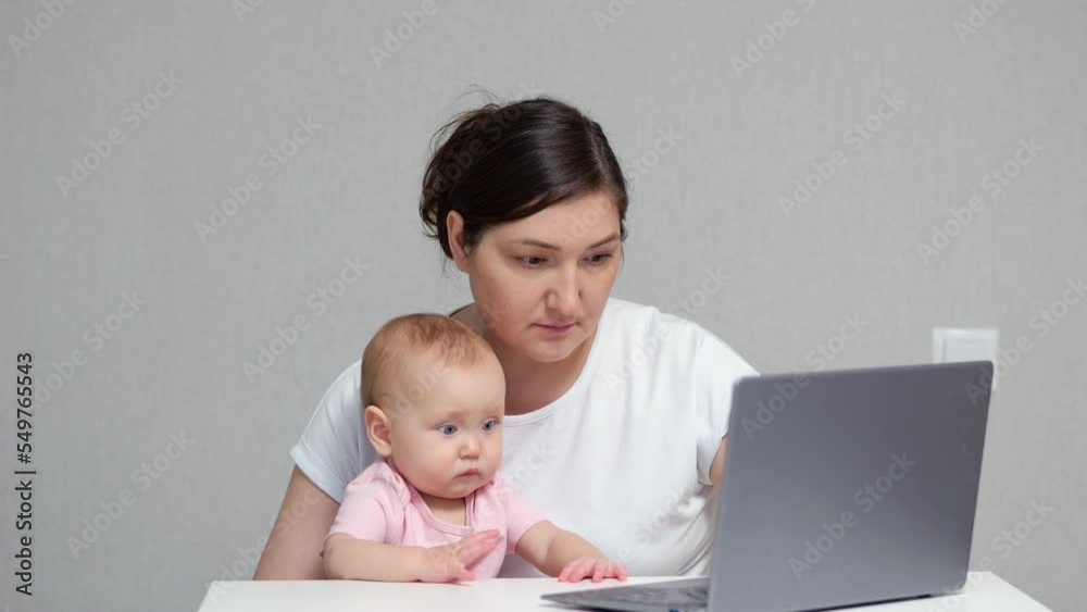 Brown-haired freelancer works on laptop holding baby daughter on lap against grey wall. Baby girl looks at monitor with confused and puzzled expression Brown-haired freelancer works on laptop holding baby daughter on lap against grey wall. Baby girl looks at monitor with confused and puzzled expression
