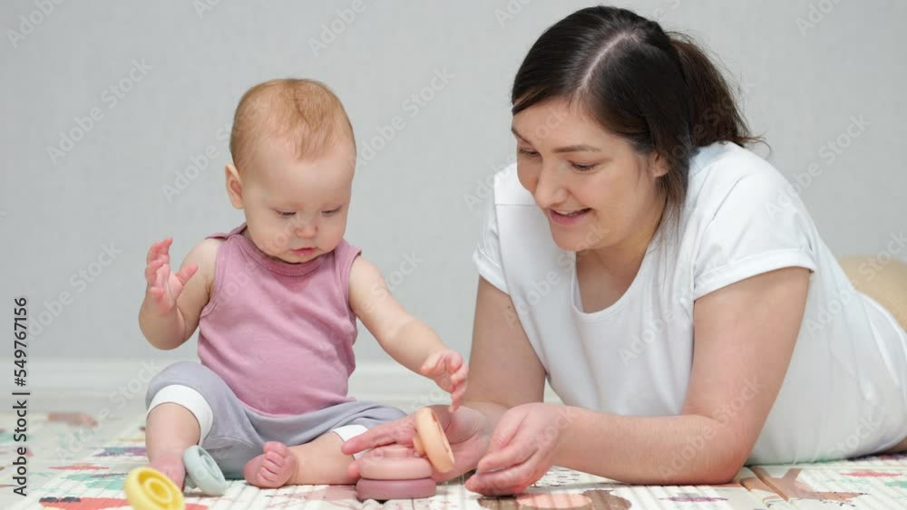 Baby girl enjoys playing pyramid toy with mother on floor. Brown-haired woman teaches excited daughter to put toys in right order and praises child work Baby girl enjoys playing pyramid toy with mother on floor. Brown-haired woman teaches excited daughter to put toys in right order and praises child work