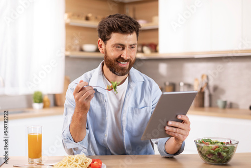 Glad adult caucasian man with beard eats lunch  read news at tablet  watch video in modern kitchen interior