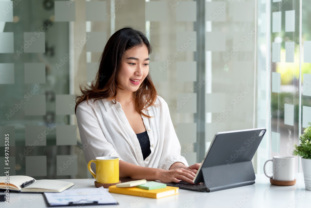 Attractive Asian business woman sitting at work happily working using tablet at office and smiling comfortably. Attractive Asian business woman sitting at work happily working using tablet at office and smiling comfortably.