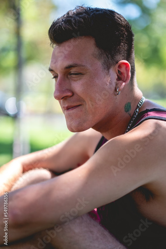 Portrait of young attractive model guy with 4-leaf clover tattoo, tank top and chains and earrings in a park in the afternoon