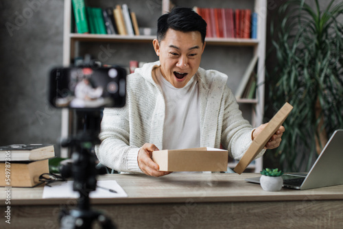 Attractive young asian man recording video using camera on smart phone while unpacking gift box. Male blogger sharing his emotions with his subscribers in social networks