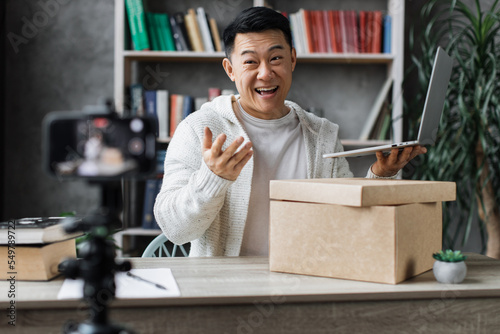 Asian man recording video on phone camera while unpacking box with new wireless laptop. Male influencer sharing with subscribers his positive feedback about new order.