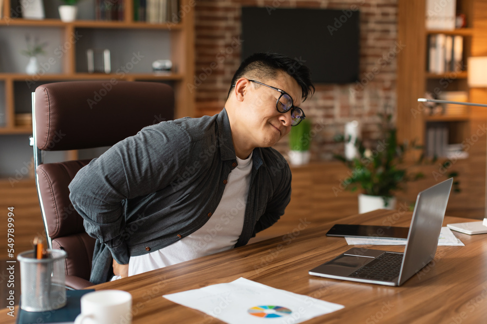 Unhappy middle aged asian man manager in glasses suffering from muscles back pain at table with laptop Unhappy middle aged asian man manager in glasses suffering from muscles back pain at table with laptop