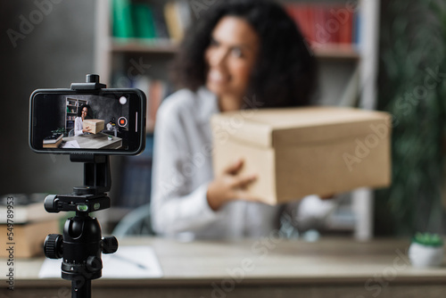 Focus on smart phone screen pretty multinational woman in casual wear recording video on camera while unpacking gift box. Female blogger sharing her emotions with her subscribers in social networks.