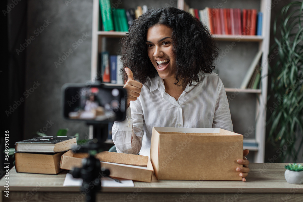 Happy female blogger unpacking gift boxes presents from companies and doing live streaming showing thumb up. Multinational woman using smart phone camera for creating content.