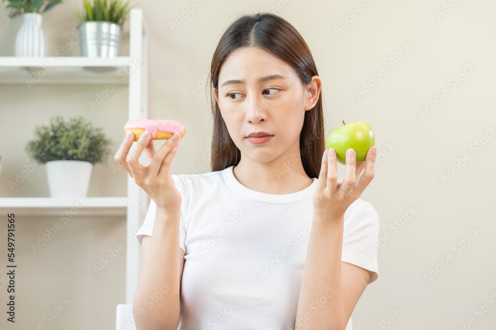 Diet, Dieting asian young woman or girl smile, confused choose, choice green apple or pink sweet donut, at home, eat food for good healthy, health when hungry. Close up female weight loss person. Diet, Dieting asian young woman or girl smile, confused choose, choice green apple or pink sweet donut, at home, eat food for good healthy, health when hungry. Close up female weight loss person.