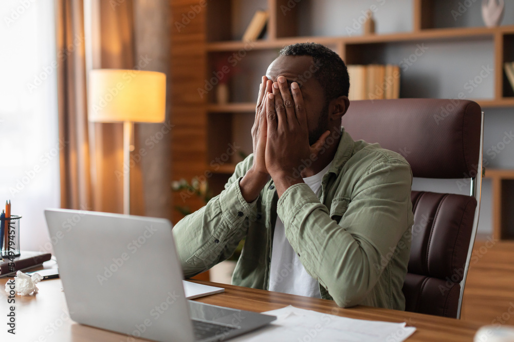 Tired sad middle aged african american guy covers face with hands suffering from overwork