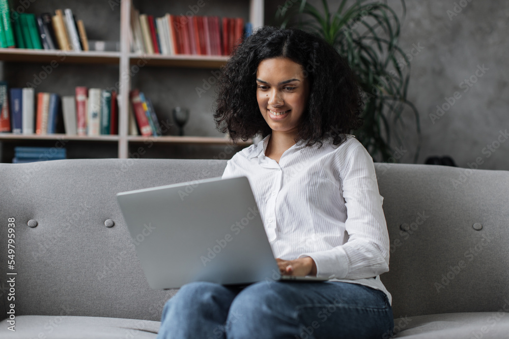 Attractive african american woman in casual clothes sitting on couch and using wireless laptop. Young female browsing internet during leisure time at cozy home. Attractive african american woman in casual clothes sitting on couch and using wireless laptop. Young female browsing internet during leisure time at cozy home.