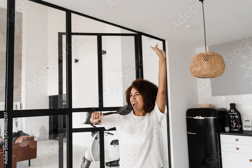 Joyful African young woman having fun with kitchen utensils. African american housewife in apron singing with spatulas for cooking microphone on the kitchen.