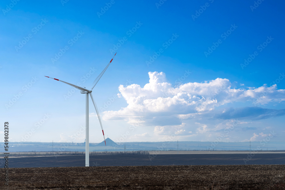 Transition to green energy. A wind turbine on the background of a light blue sky with a white cloud. In the background is a coal mine with a waste rock dump.