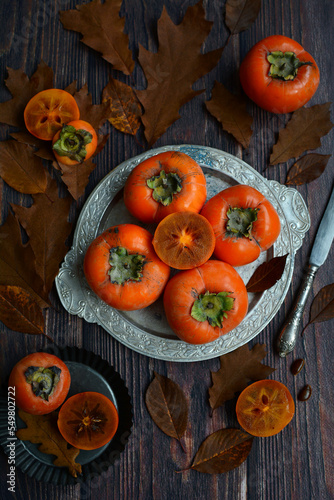 Vertical composition. Ripe appetizing persimmon fruits on a plate on a dark wooden background. Autumn fruits
