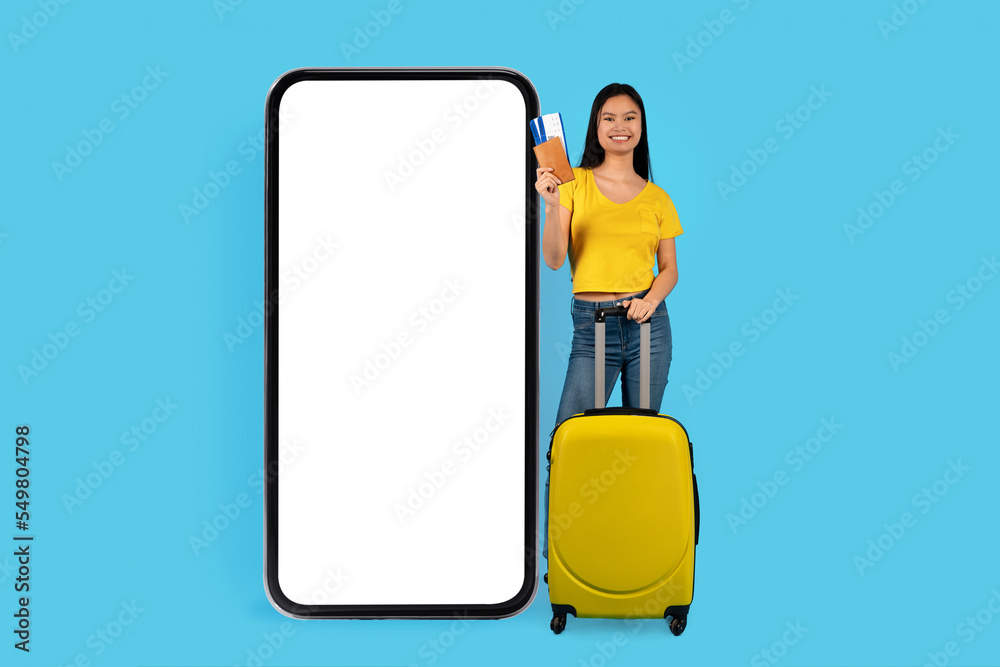 Smiling millennial japanese lady blogger with suitcase shows passport and tickets, near huge smartphone Smiling millennial japanese lady blogger with suitcase shows passport and tickets, near huge smartphone