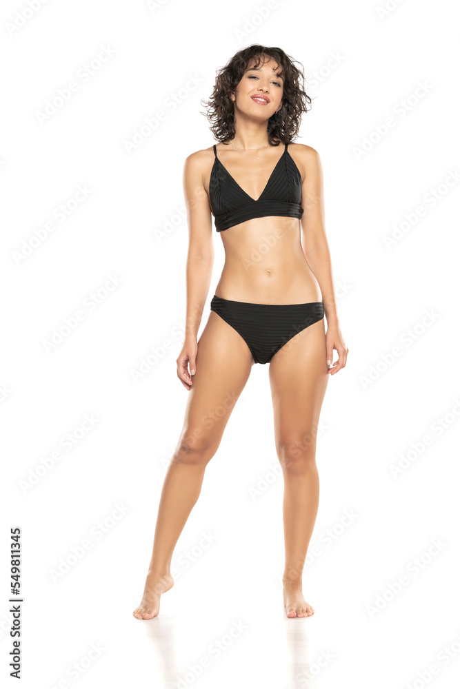 Young smiling exotic brunette woman in black bikini swimsuit posing on a white studio background. Front view. Young smiling exotic brunette woman in black bikini swimsuit posing on a white studio background. Front view.
