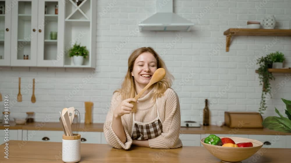 A woman is cooking in kitchen and having fun. The housewife holds wooden spatula in her hand and swings it and laughs and looks at spoon. Portrait of smiling girl. Having fun preparing food. Housework A woman is cooking in kitchen and having fun. The housewife holds wooden spatula in her hand and swings it and laughs and looks at spoon. Portrait of smiling girl. Having fun preparing food. Housework