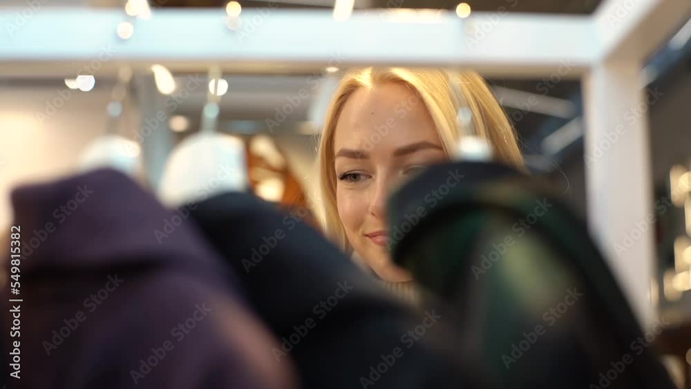 Close-up face of pretty blonde woman with charming smile plucked hanger choosing clothes in clothing store. Concept of sale promotion, consumerism and shopping. Shooting in slow motion. Close-up face of pretty blonde woman with charming smile plucked hanger choosing clothes in clothing store. Concept of sale promotion, consumerism and shopping. Shooting in slow motion.