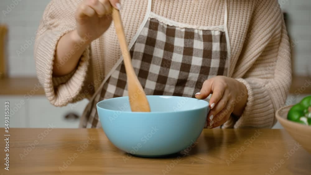 The cook prepares food and mixes ingredients in deep bowl with wooden spatula. Healthy Eating. Homemade dishes. A housewife in apron prepares dinner in kitchen. Eco tools. Kitchen tools and utensils