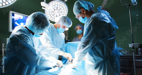 Group of professional mixed-races doctors in uniform performing neurosurgery operation under bright lamps using medical instruments and items in dark operating room in hospital. Surgery concept photo