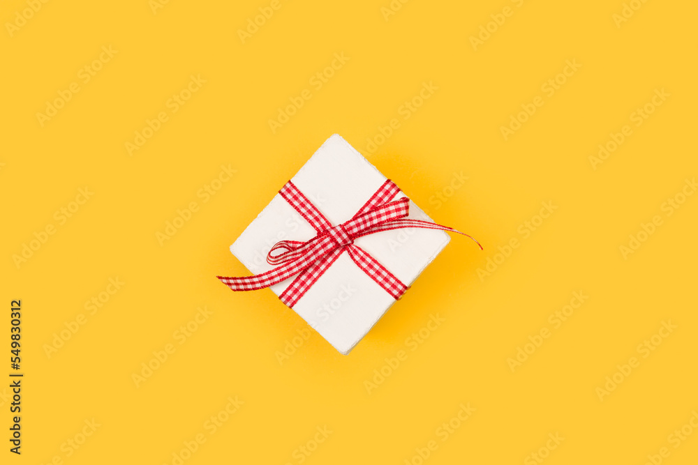 Fototapeta premium Caja de regalo blanca con un lazo de color rojo y blanco sobre un fondo amarillo liso y aislado. Vista superior y de cerca. Copy space