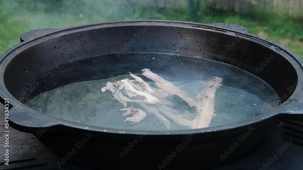 A cook in black gloves puts pieces of raw fish into a vat of boiling water for cooking fish soup. A cook in black gloves puts pieces of raw fish into a vat of boiling water for cooking fish soup.