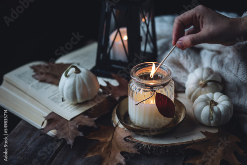 Young woman in the knitted warm sweater lights a candle on a wooden table. Cozy fall atmosphere  beautiful autumn home decor with pumpkins and leaves
