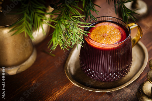 Mulled wine with cinnamon and orange served in festive Christmas decoration on wooden board. Party and New Year atmosphere. Christmas tree in background, a mug with Christmas motive. 