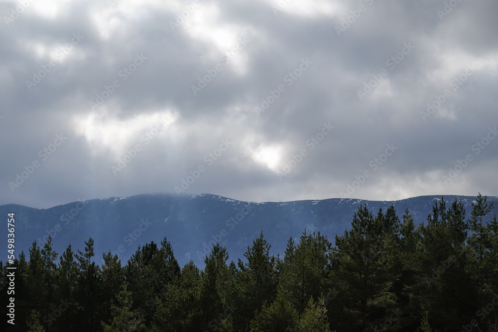Sunrays falling between the pine tree forest and snow covered mountains through the murky clouds Sunrays falling between the pine tree forest and snow covered mountains through the murky clouds