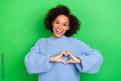 Portrait of toothy beaming stylish girl with wavy coiffure wear blue oversize pullover showing heart isolated on green color background