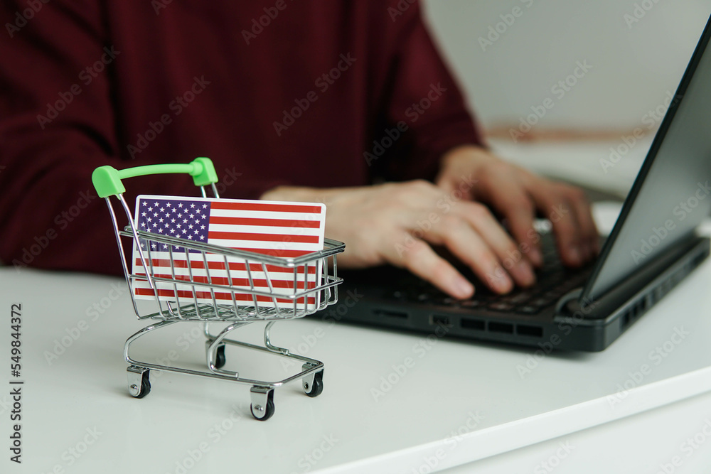 Small shopping cart with flag of the United States of America USA on the table. Man using laptop for shopping online. Small shopping cart with flag of the United States of America USA on the table. Man using laptop for shopping online.