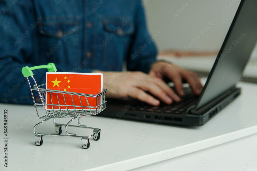 Small shopping cart with chinese flag on the table. Man using laptop for shopping online.