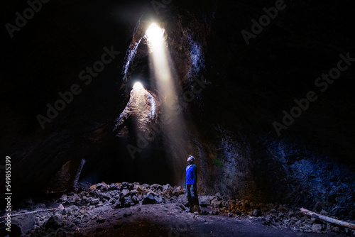 Adventurous athletic male standing in a lava tube looking at the sunlight shinning down into the cave through holes in the ceiling.
