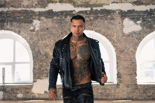 hot tattooed man in leather against the big window.Handsome Young Athletic Male Fashion Model.Muscular athletic sexy male with naked torso. Brutal handsome man with tattooed body.